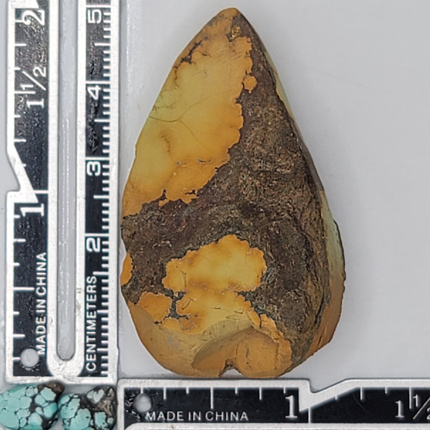 A natural turquoise stone weighing 24 grams, featuring earthy golden hues with bold dark matrix patterns and organic textures. The stone measures approximately 5.5 cm x 3 cm x 0.6 cm, displayed with a ruler for scale. Available for purchase at www.naturalturquoise.com, ideal for lapidary and jewelry projects.